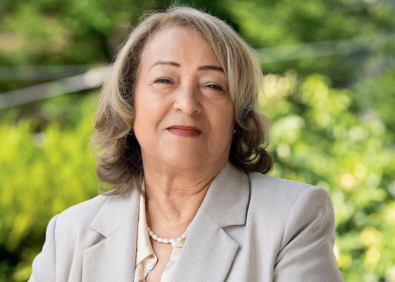 Une femme plus âgée aux cheveux gris mi-longs, vêtue d'un blazer gris clair et d'un chemisier blanc, se tient dehors avec de la verdure en arrière-plan - un moment serein capturé dans le cadre de son Témoignage pour Femina.